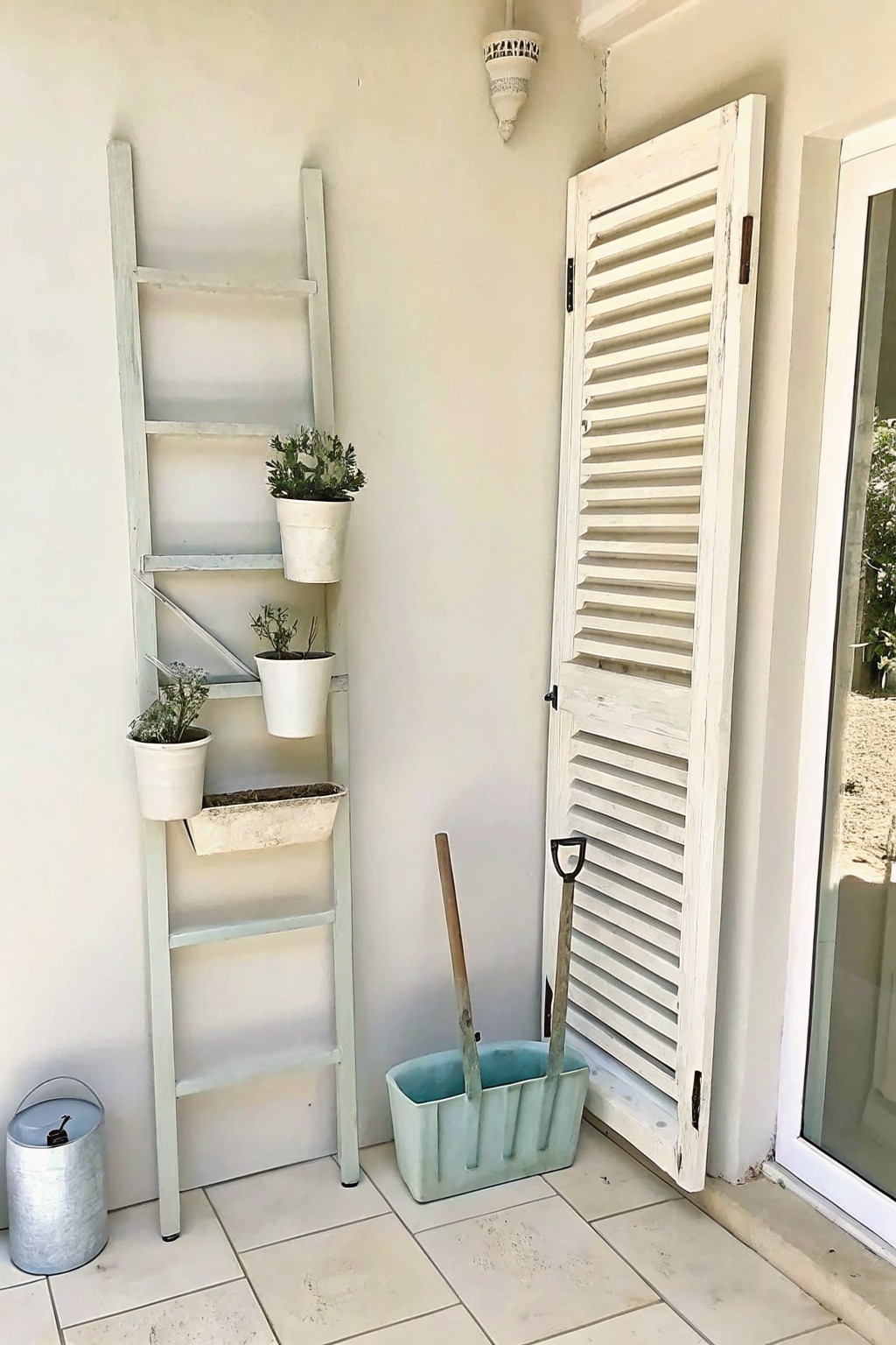A light green wooden ladder leans against a wall on a tiled porch, holding several white pots with plants beside a white shutter door and a light blue container with garden tools.