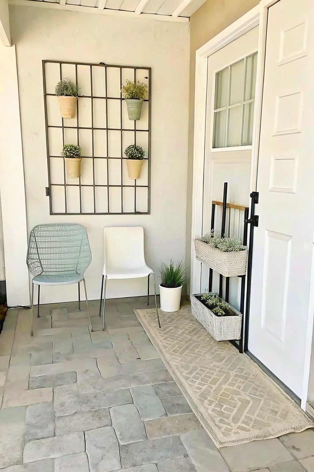 A covered porch with a large metal grid on the wall holding four small hanging pots, two chairs on stone flooring, and woven planters beside a white door.
