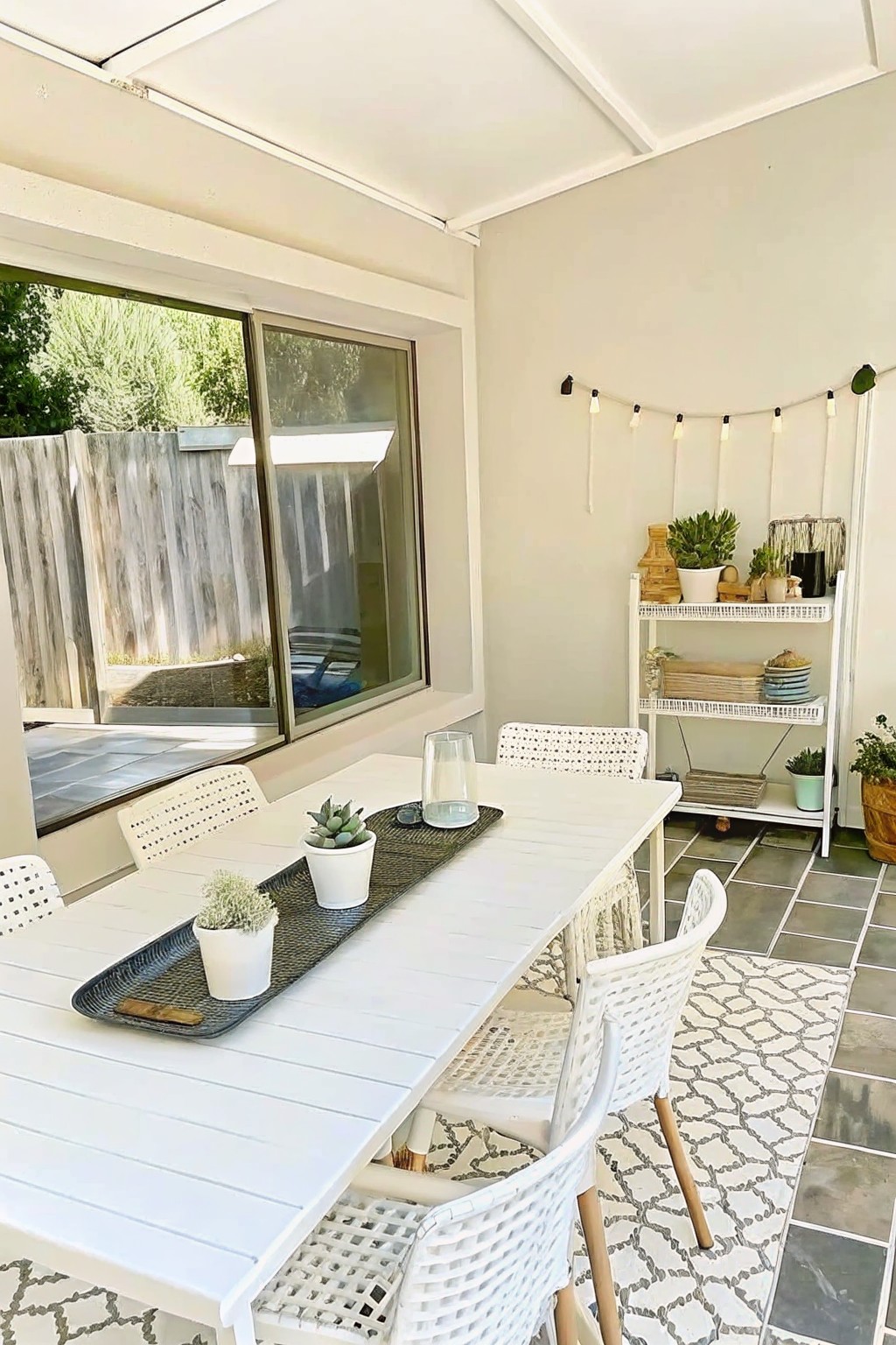 White patio dining table with woven chairs, succulents, shelves, and string lights.