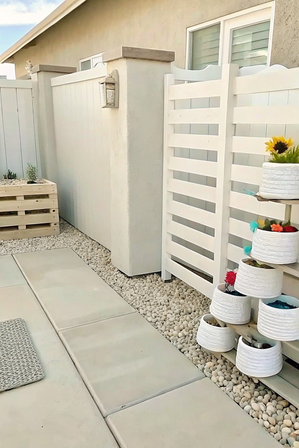 Backyard path with concrete pavers and gravel beside a white fence, showing a wooden crate used as a planter.