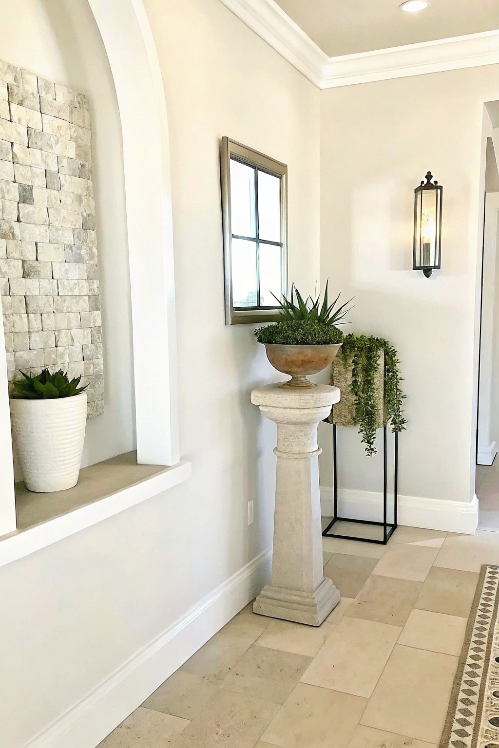 A bright indoor hallway shows a tall stone pedestal holding a large weathered bowl planter with greenery, a recessed niche with a white ribbed pot, and a black metal stand with hanging plants nearby.