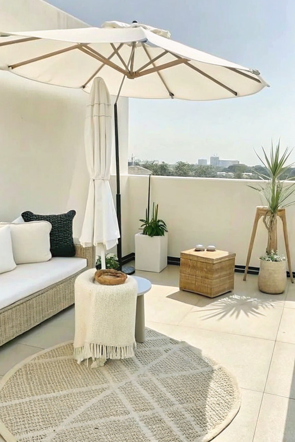 A sunlit balcony with a white sofa, large umbrella, round rug, and several potted plants of varying heights placed around the seating area.
