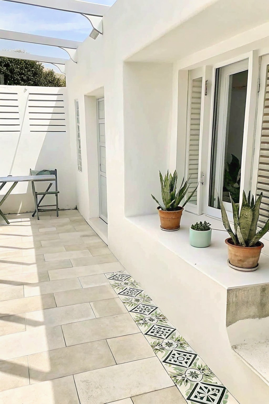 White patio with beige tiles, patterned border, potted plants, folding table and chair.