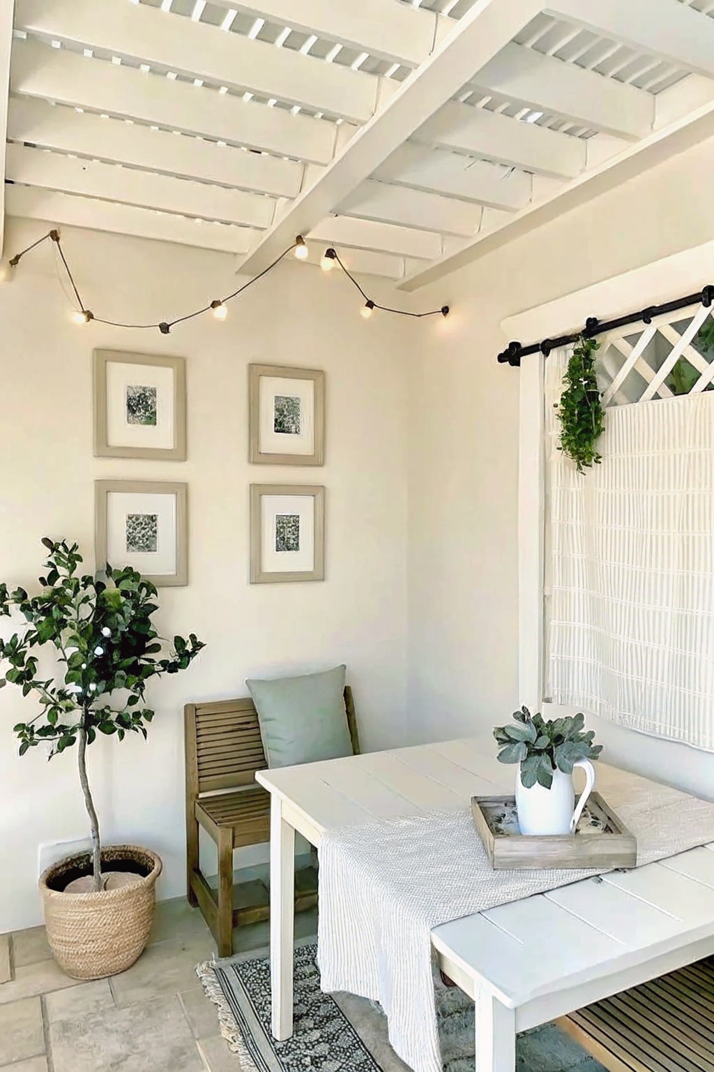An outdoor patio corner with a white wooden table and bench, a large potted tree in a woven basket on the floor, a small plant in a white pitcher on a tray, string lights overhead, and framed pictures on the wall.