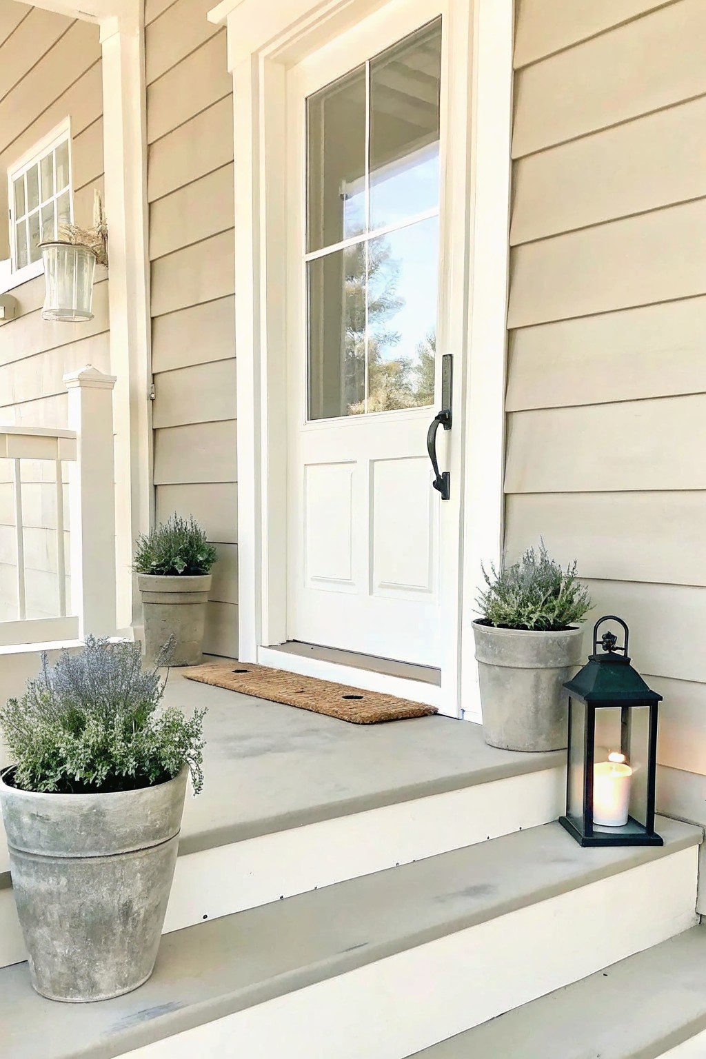 Front entrance of a house with white door and siding, concrete steps holding several large gray pots with plants, a woven doormat, and a black lantern with a lit candle.