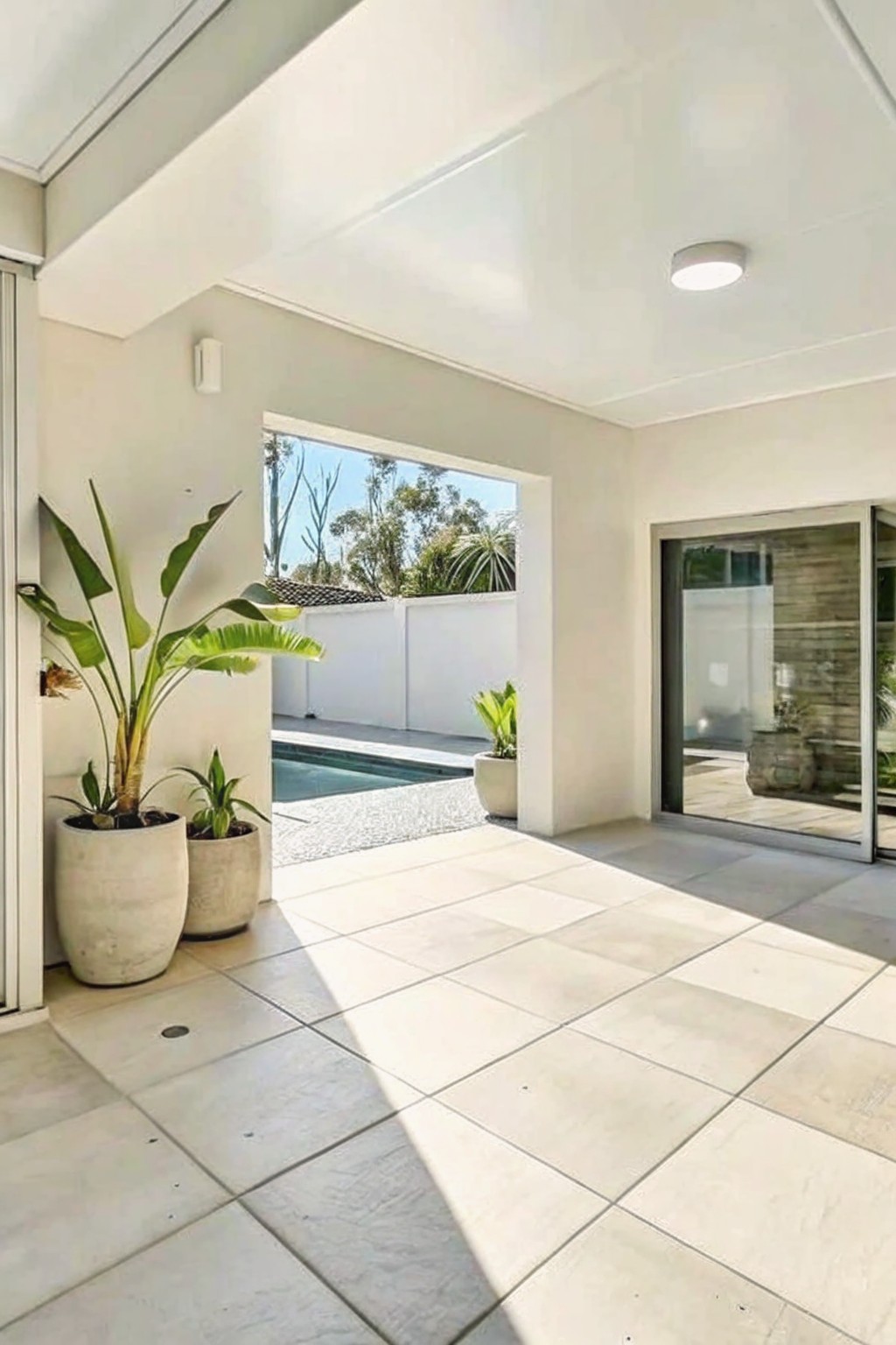 A covered outdoor patio with large potted plants clustered on the left near a doorway that opens to a pool area.