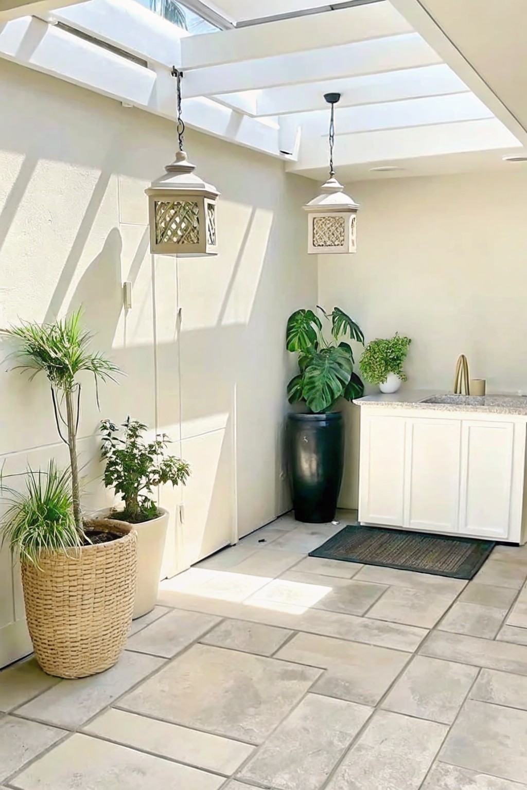 An outdoor patio with stone tile flooring, white walls, hanging lanterns, a white cabinet with sink, and several potted plants including one in a large woven basket and one in a tall dark container.