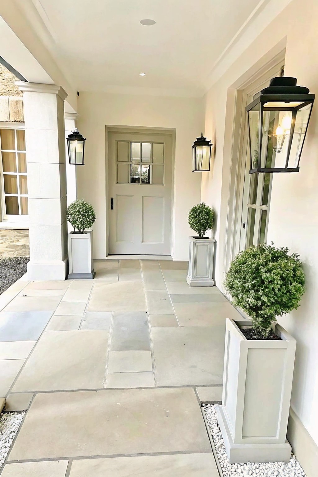 A front entry with a white door centered between two matching white square planters holding rounded green shrubs, black wall lanterns on either side, and a light stone tile walkway.