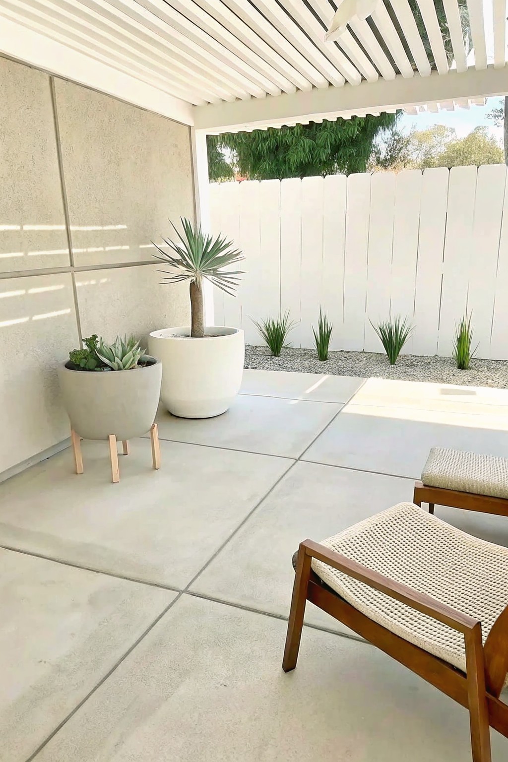 Outdoor patio with several large concrete pots holding plants of varying heights next to woven chairs and a white fence.