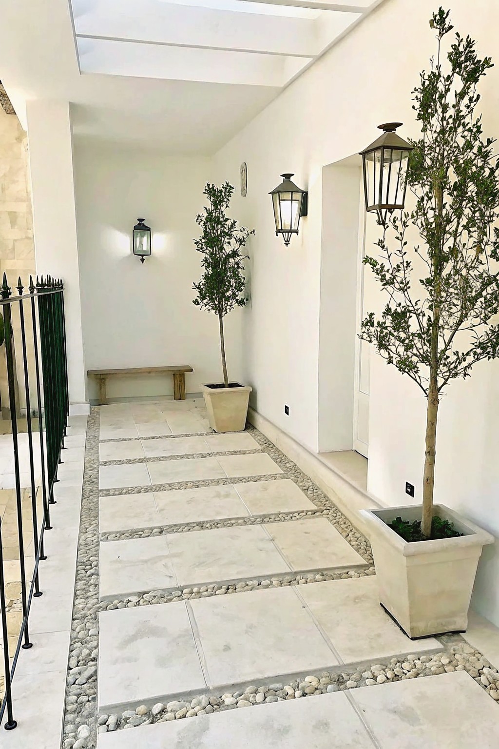 A covered outdoor walkway with large square planters holding tall trees placed beside a stone path bordered by pebbles and lit by wall lanterns.