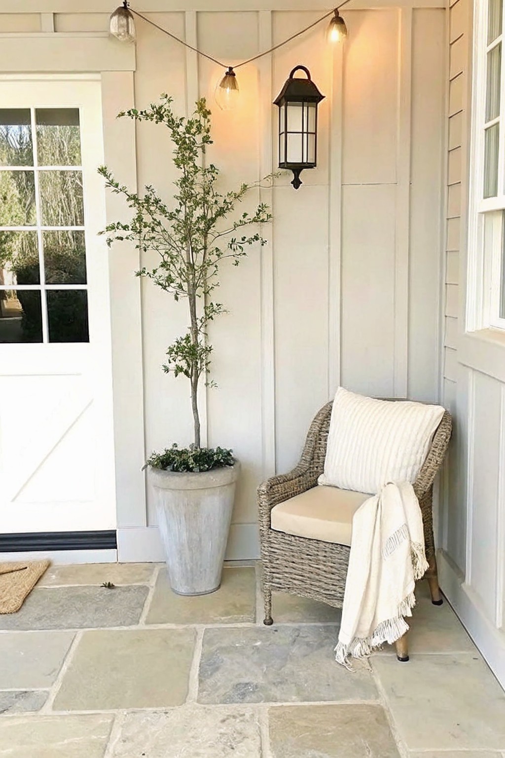 A tall tree in a large gray pot stands beside a wicker chair with a cushion and throw on a stone porch.