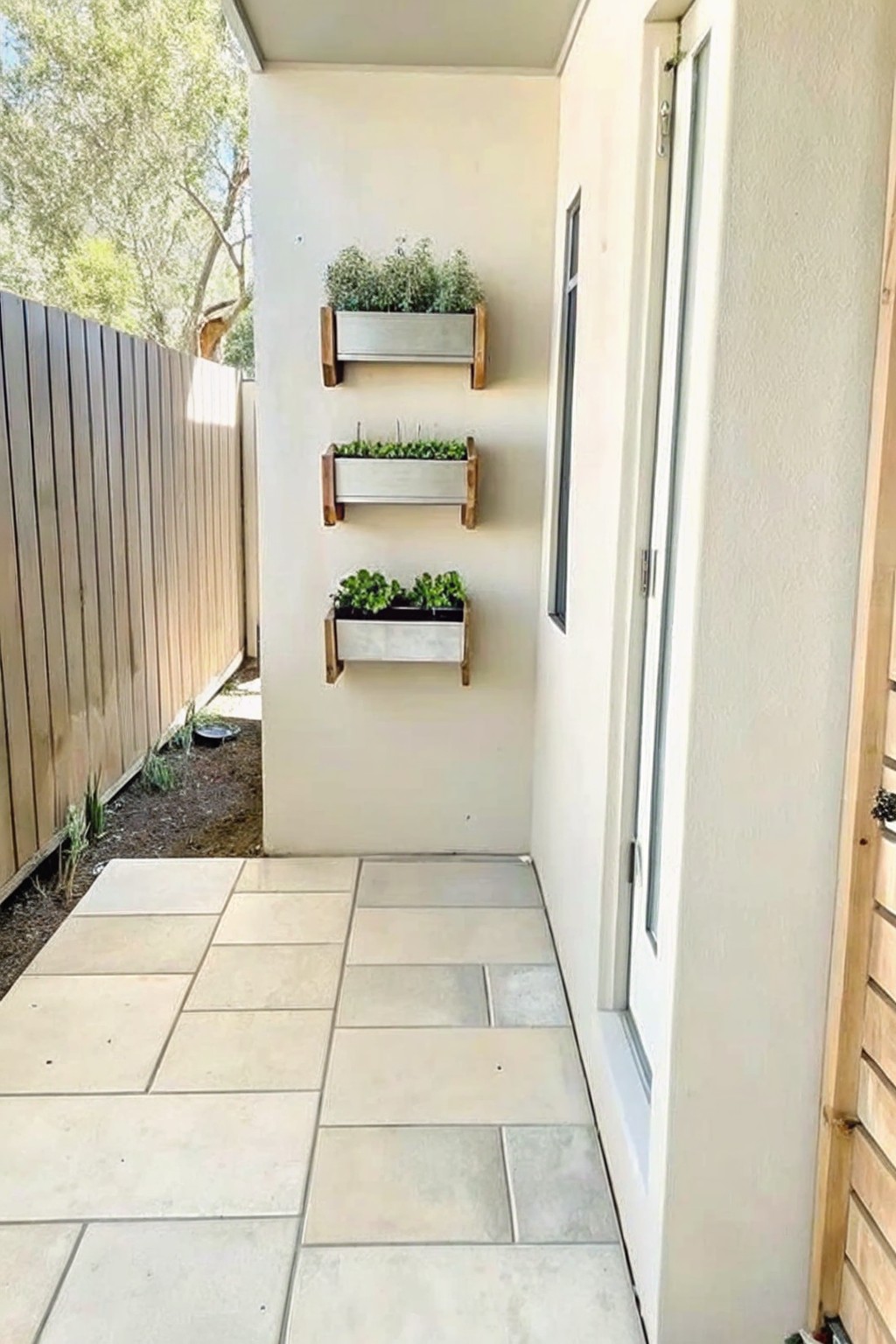 Three rectangular wall-mounted planters with green plants on a beige exterior wall next to a paved walkway and white door.