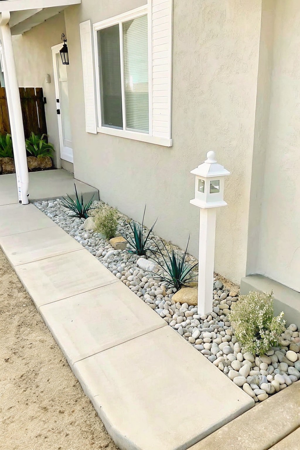 A narrow gravel bed with small white and gray stones runs along a concrete walkway beside a beige stucco house wall, with several spiky green plants and a white lamp post.
