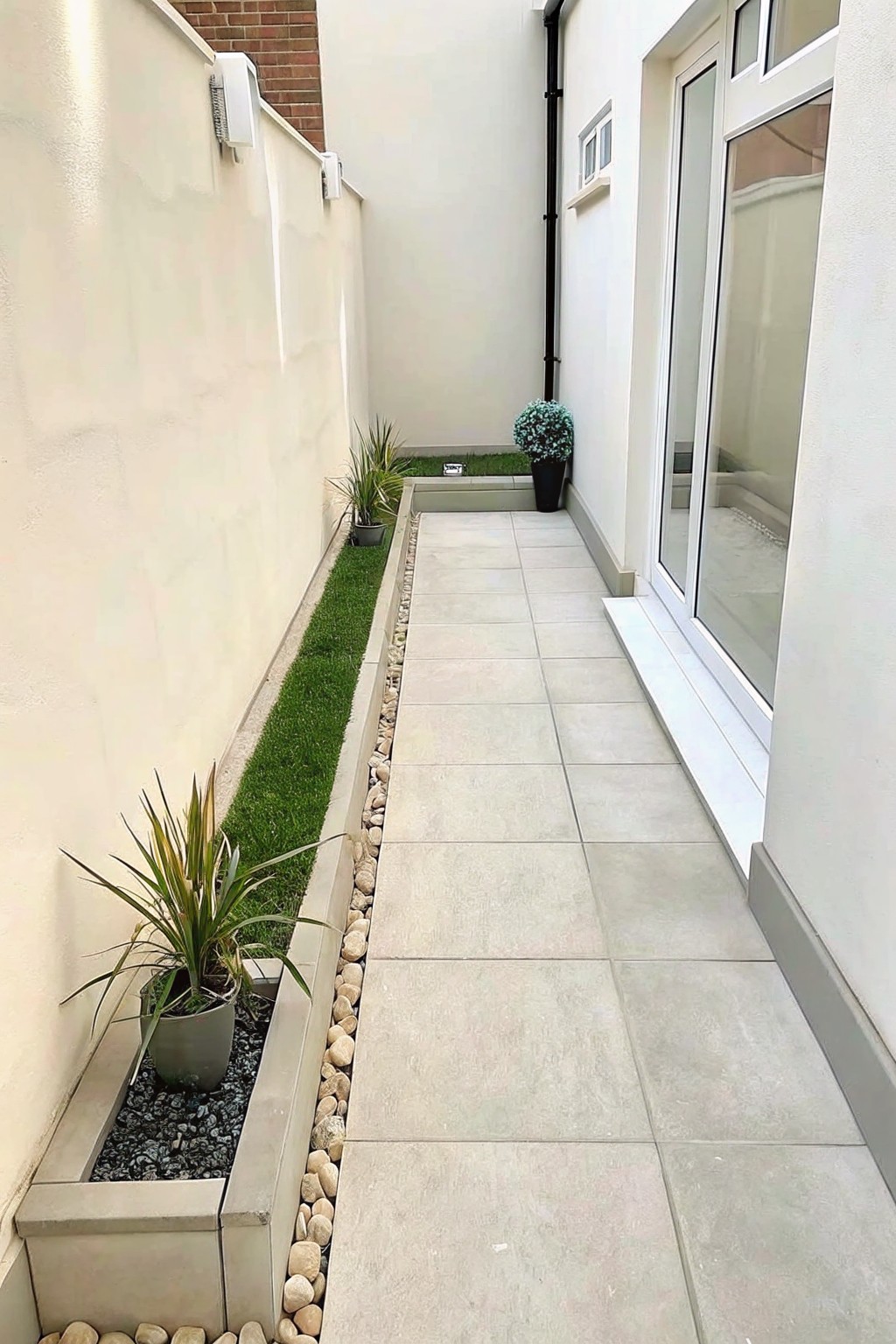 A narrow paved side path bordered by gravel strips and low planting beds with potted plants along a white wall.