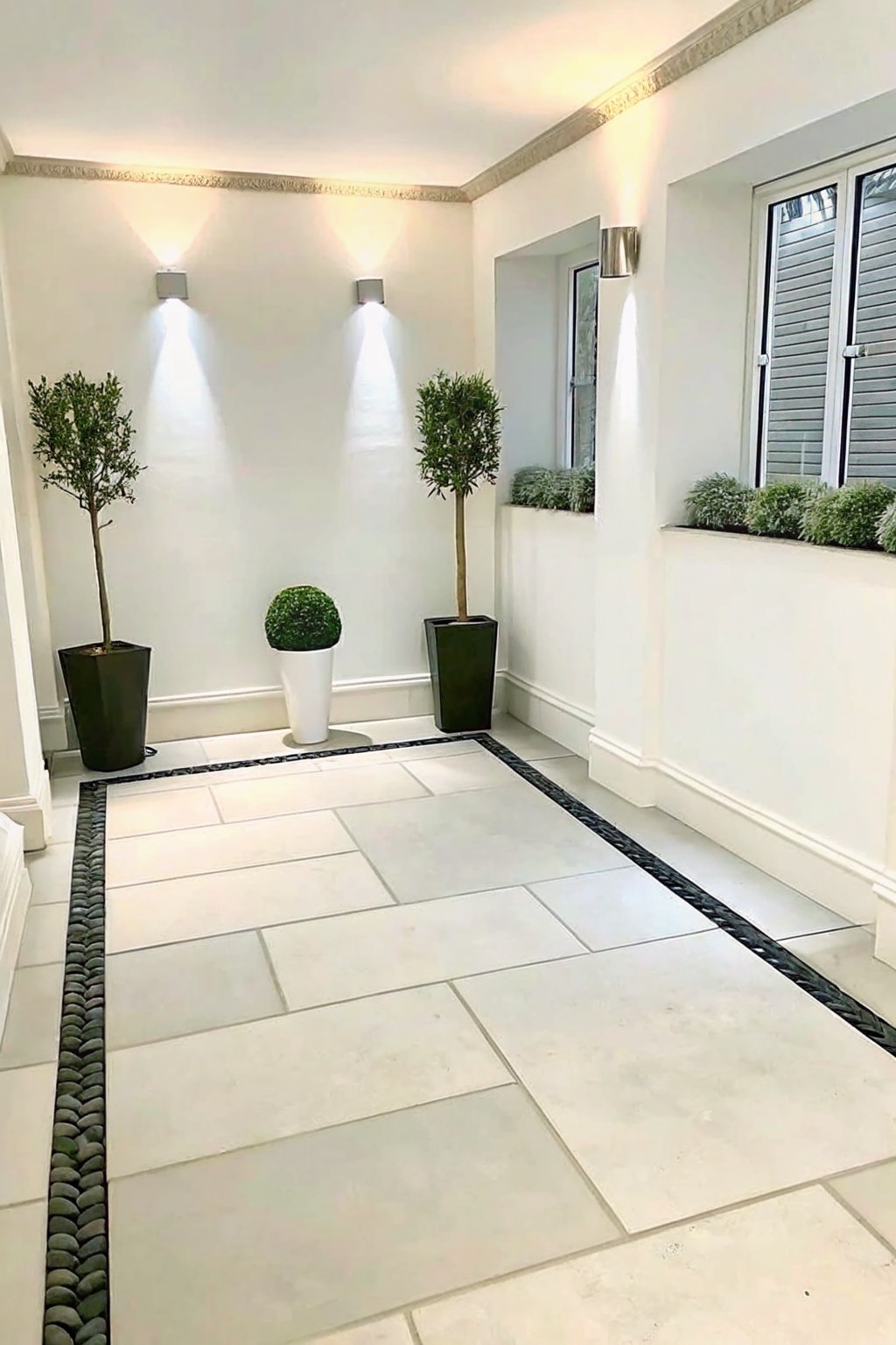 A light-colored stone tile walkway bordered on both sides by narrow lines of dark rounded pebbles, with potted plants placed along the edges against white walls.