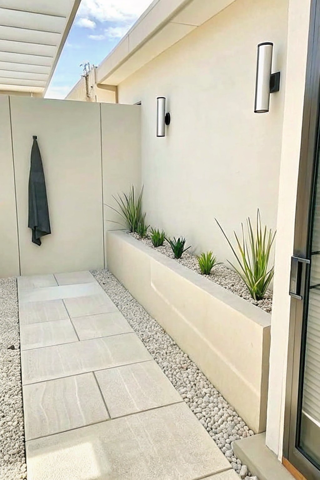A narrow paved walkway with large light tiles runs beside a long raised planter filled with white gravel and scattered green plants, set against a plain wall with two wall sconces.