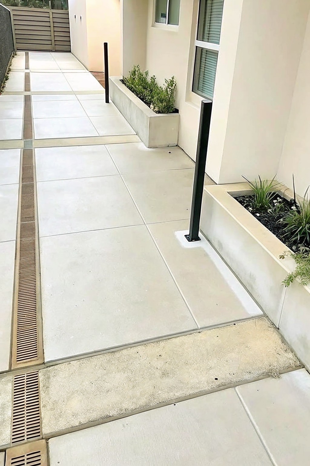 A wide concrete paver walkway with a linear drain grate on the left, bordered by raised rectangular concrete planters holding green plants and black metal posts.