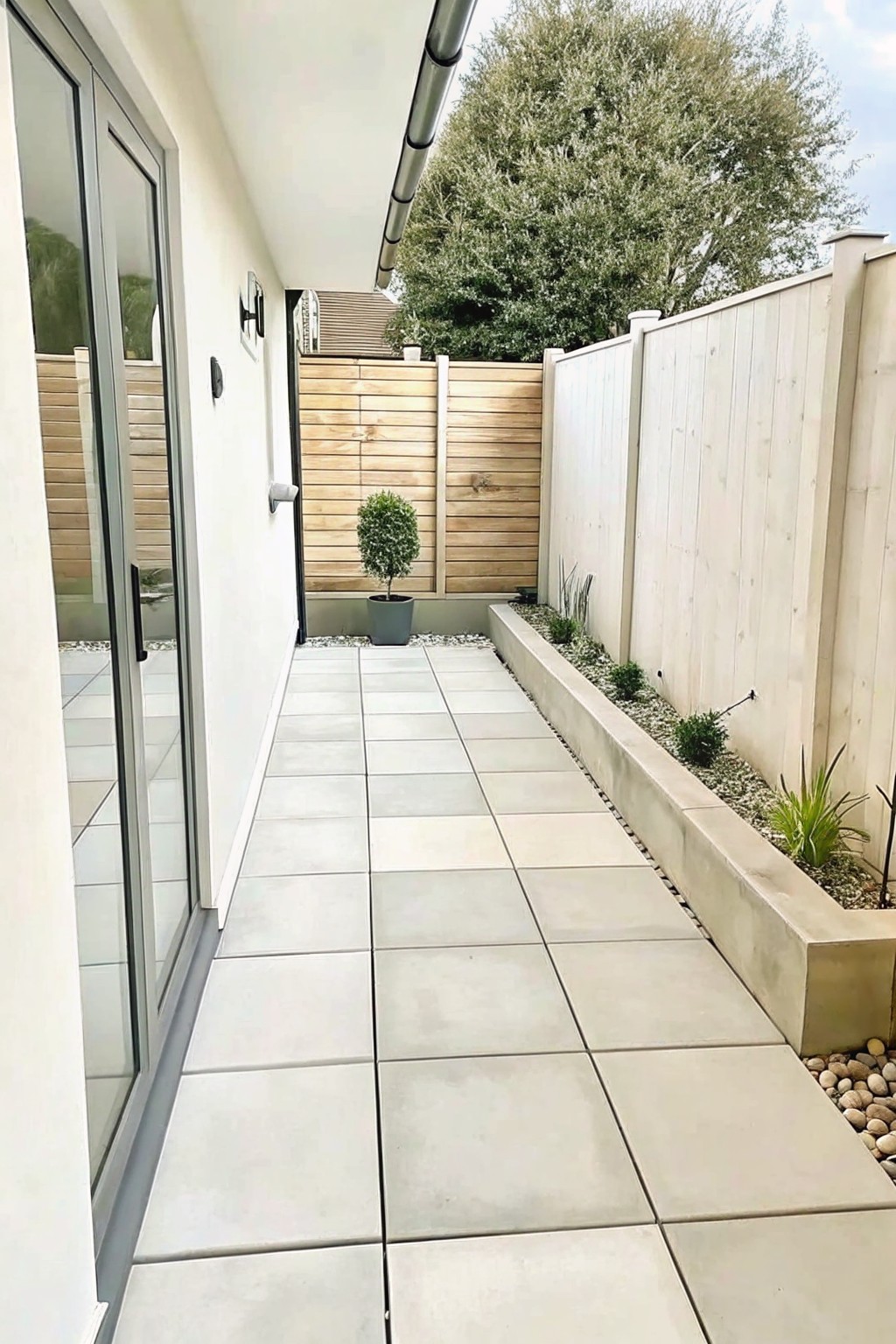 Narrow tiled walkway with glass doors, wooden fence, and concrete planter bed