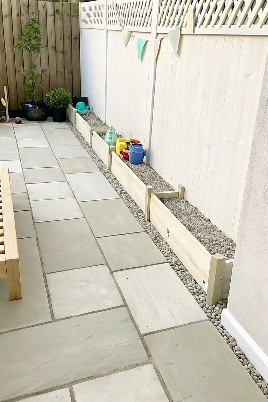 Backyard patio with large gray paving stones beside light wooden raised borders filled with gravel along a white fence, with a wooden bench visible on the left.