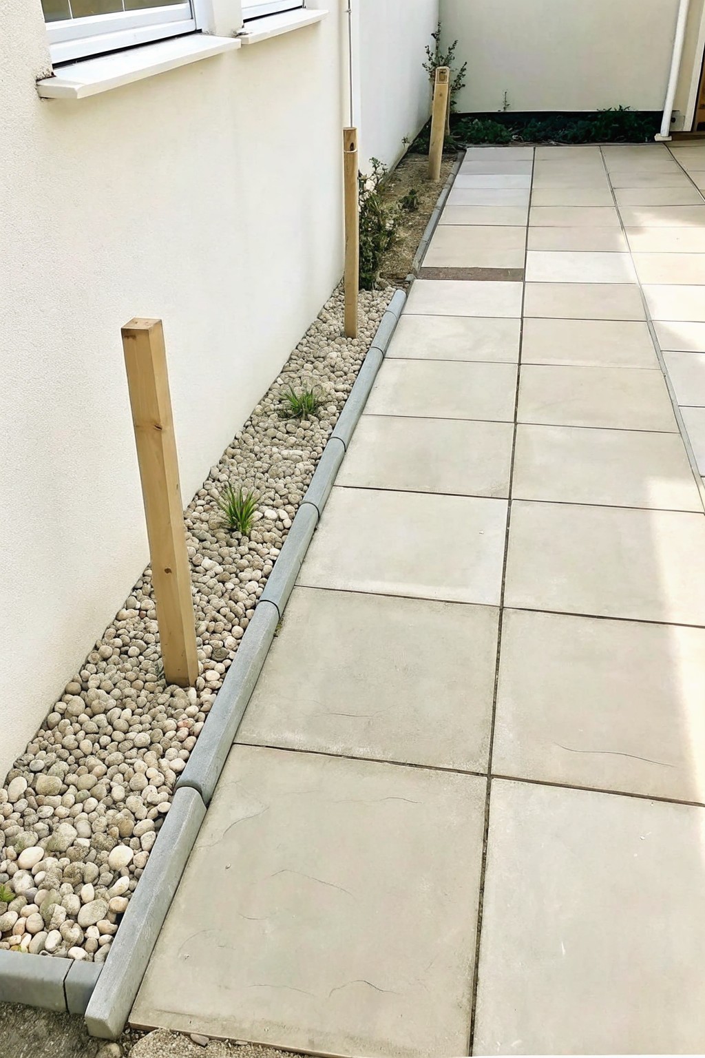 A concrete paved path runs beside a narrow gravel strip lined with wooden posts and a white wall.