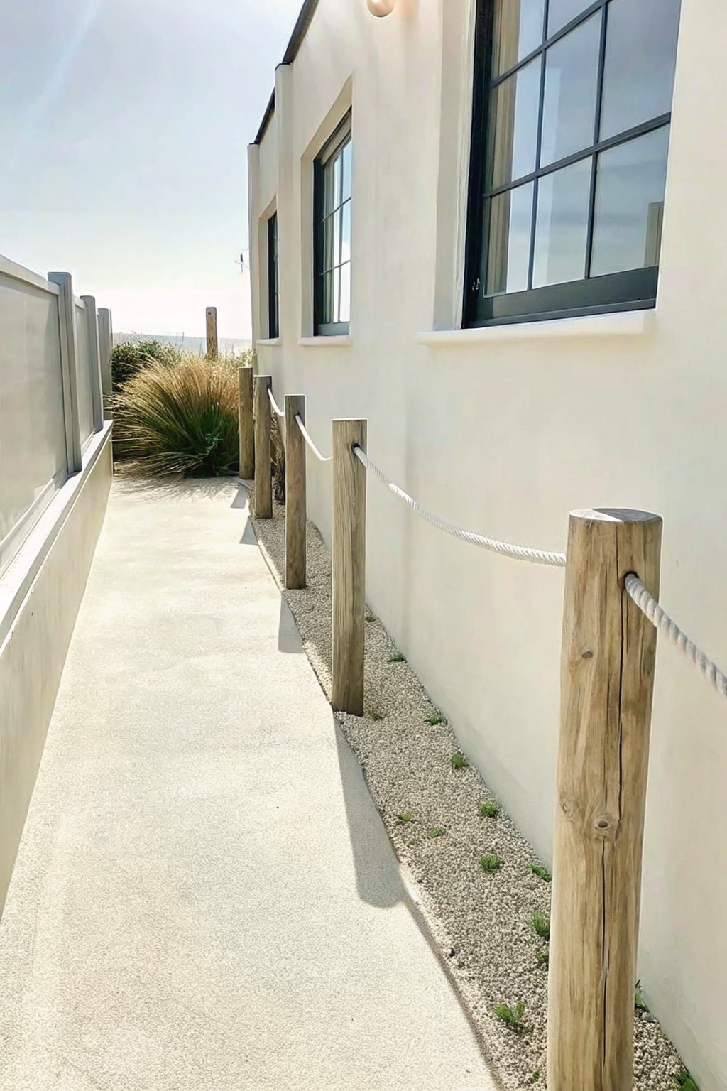 A concrete path beside a white building with a gravel border held by wooden posts and a white rope.