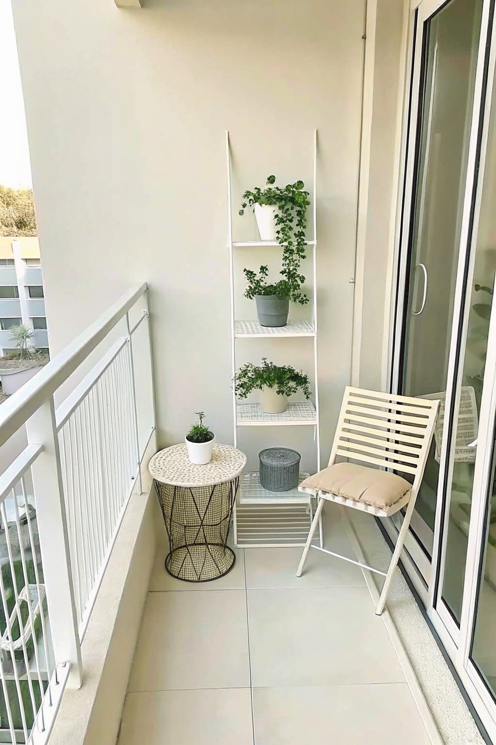 A narrow balcony with a white ladder shelf holding several potted plants, a folding chair with a cushion, and a small round table on light tiles.