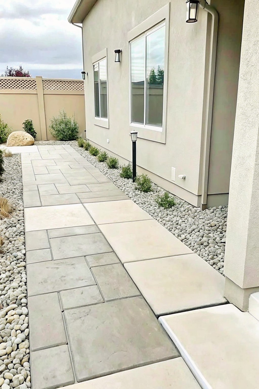 A concrete paver walkway bordered by a strip of white gravel and low shrubs runs along the side of a beige house.