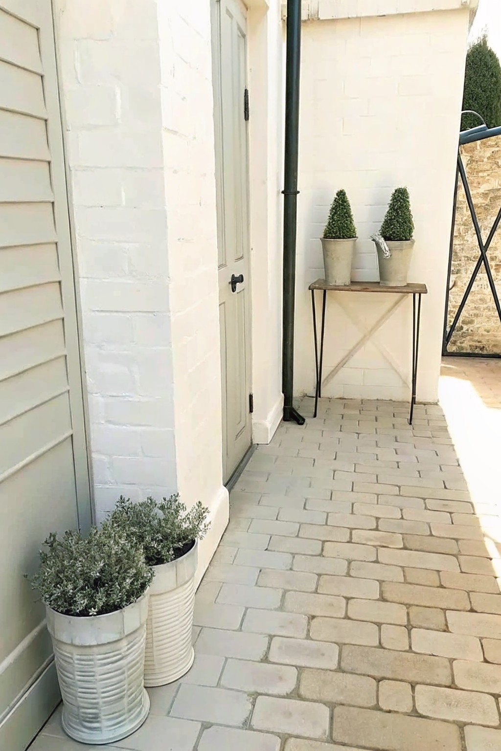 A paved outdoor walkway beside a white brick wall with two potted shrubs on a narrow console table and two larger pots on the ground near a door.