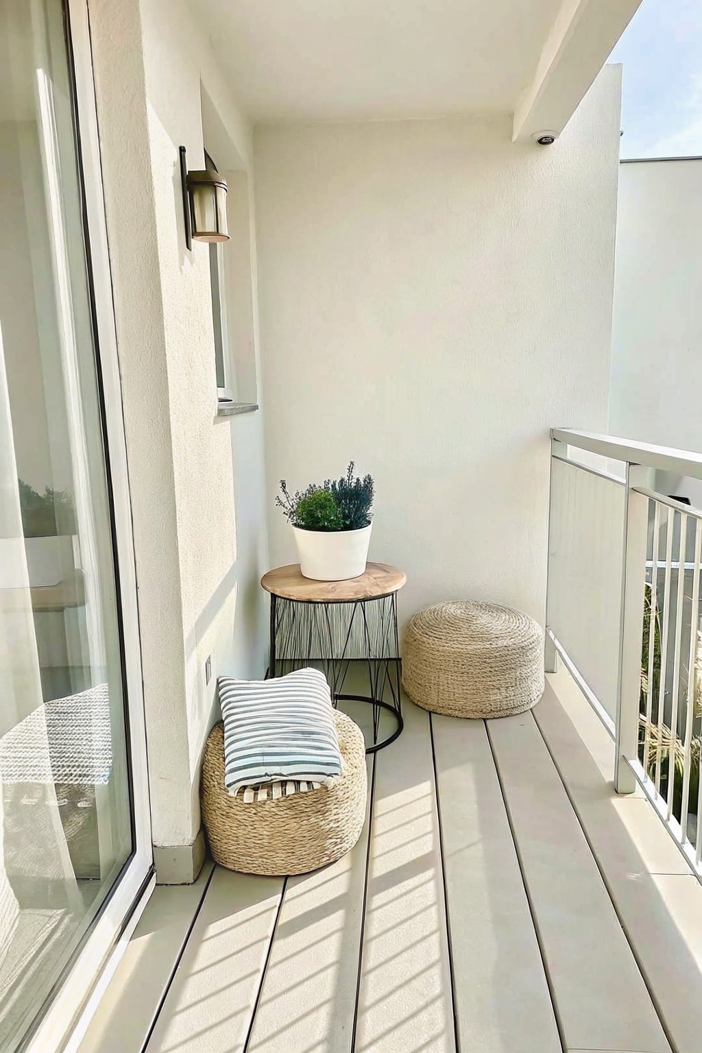 A narrow balcony shows a round wooden side table with a white potted plant, two woven poufs, and a metal railing along the right side.