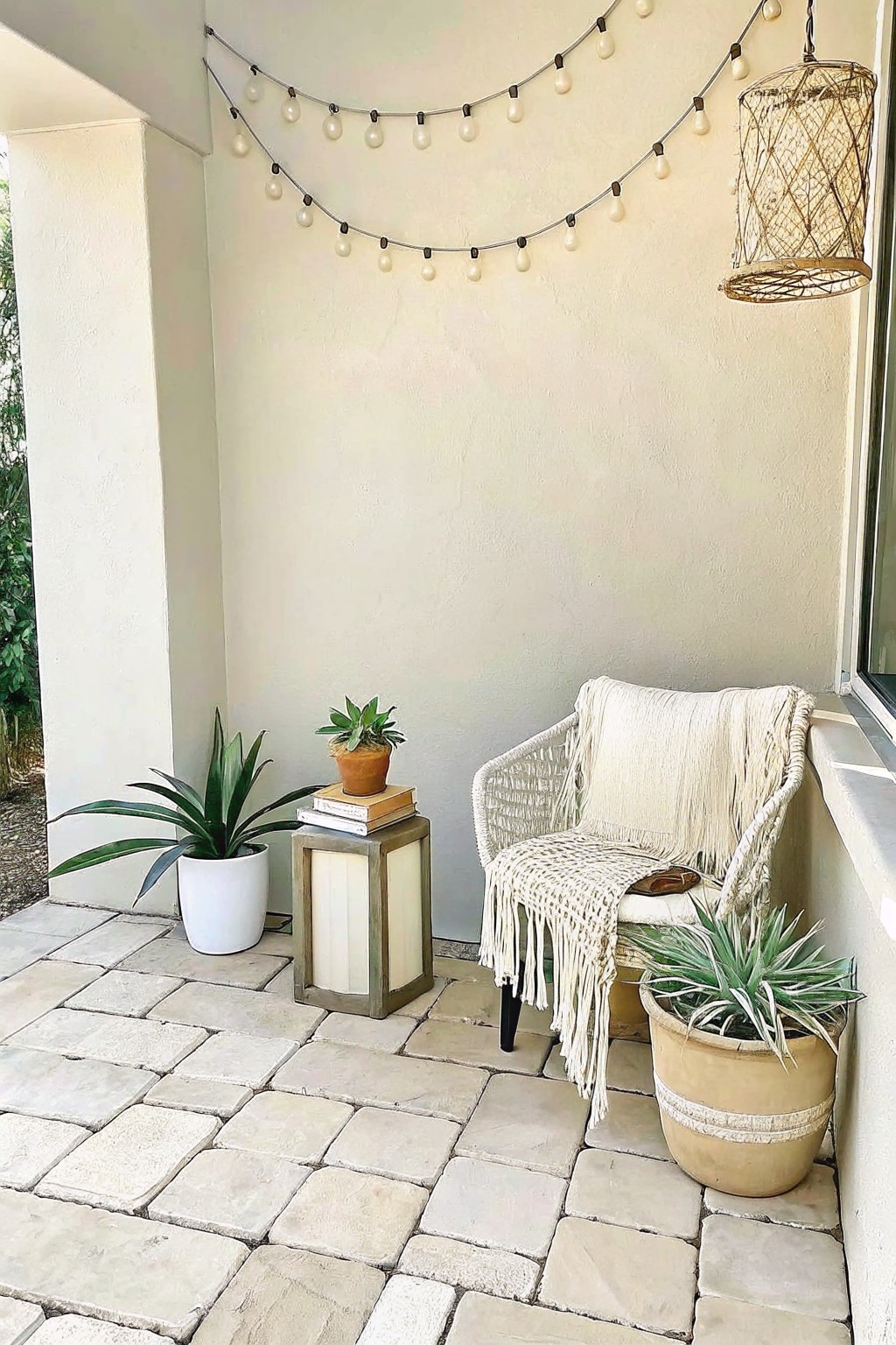 A stone patio corner shows a woven chair with throws, a small wooden side table holding books and a potted plant, and several larger potted plants arranged on the floor beneath string lights.