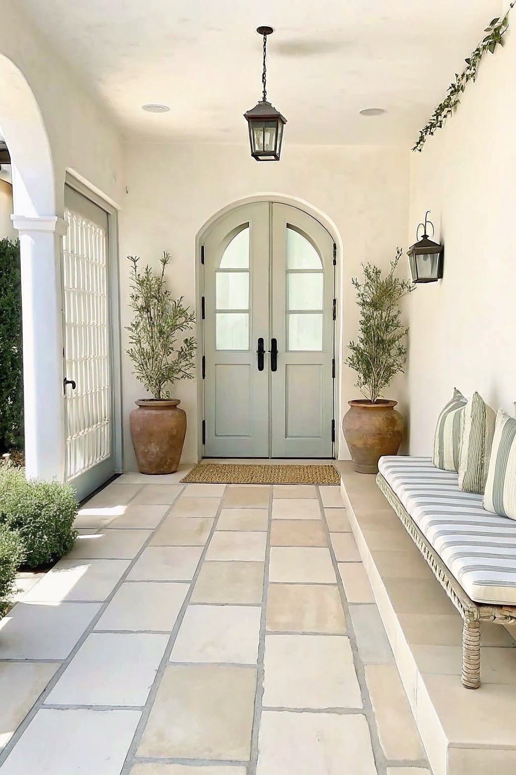 A covered patio with beige stone tiles, light green double doors under an arch, two large terracotta pots holding shrubs on either side of the doors, a striped cushion bench along the right wall, and two black lanterns.
