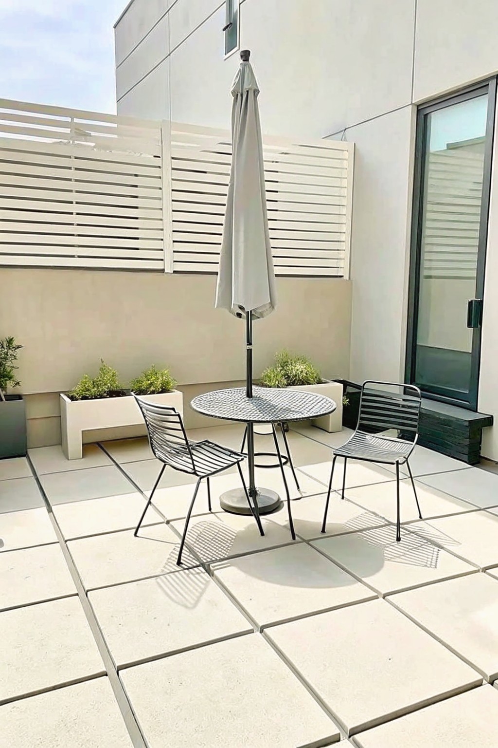 A modern patio with a round metal table under a closed umbrella, two wireframe chairs, and rectangular planters along the back wall.