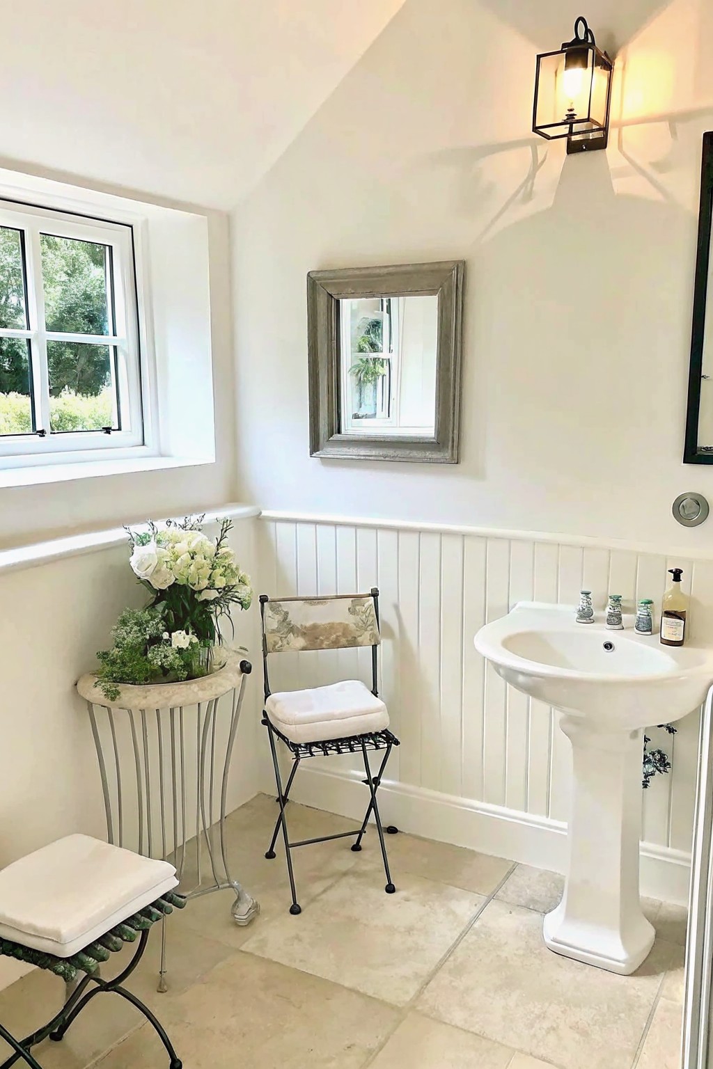 A corner of a white bathroom showing a round stand with a large floral arrangement, a folding chair with a cushion, a pedestal sink, and a window on the left wall.