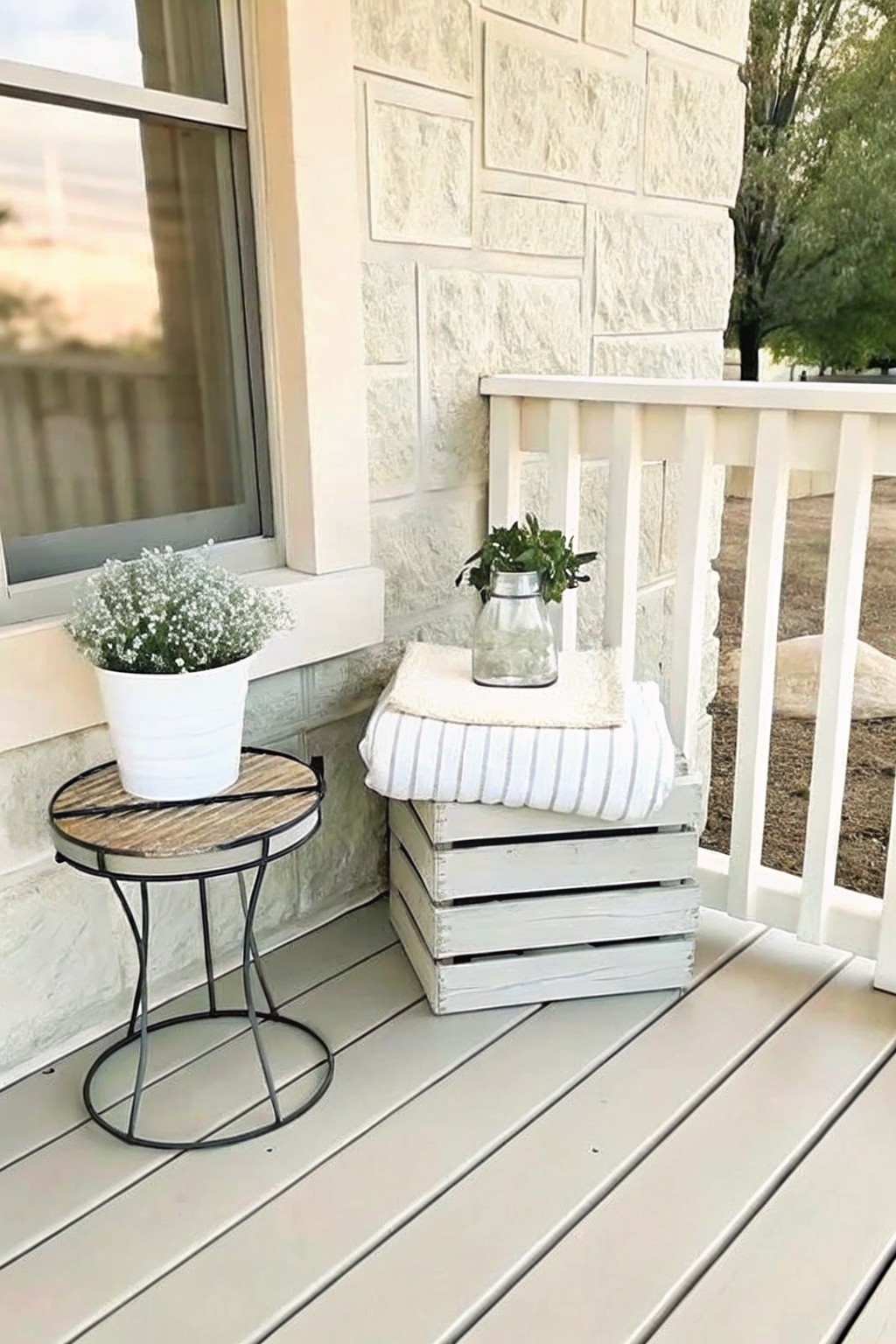 A round metal table with a white plant pot sits beside stacked white wooden crates topped with a striped towel and glass vase on a light-colored porch.