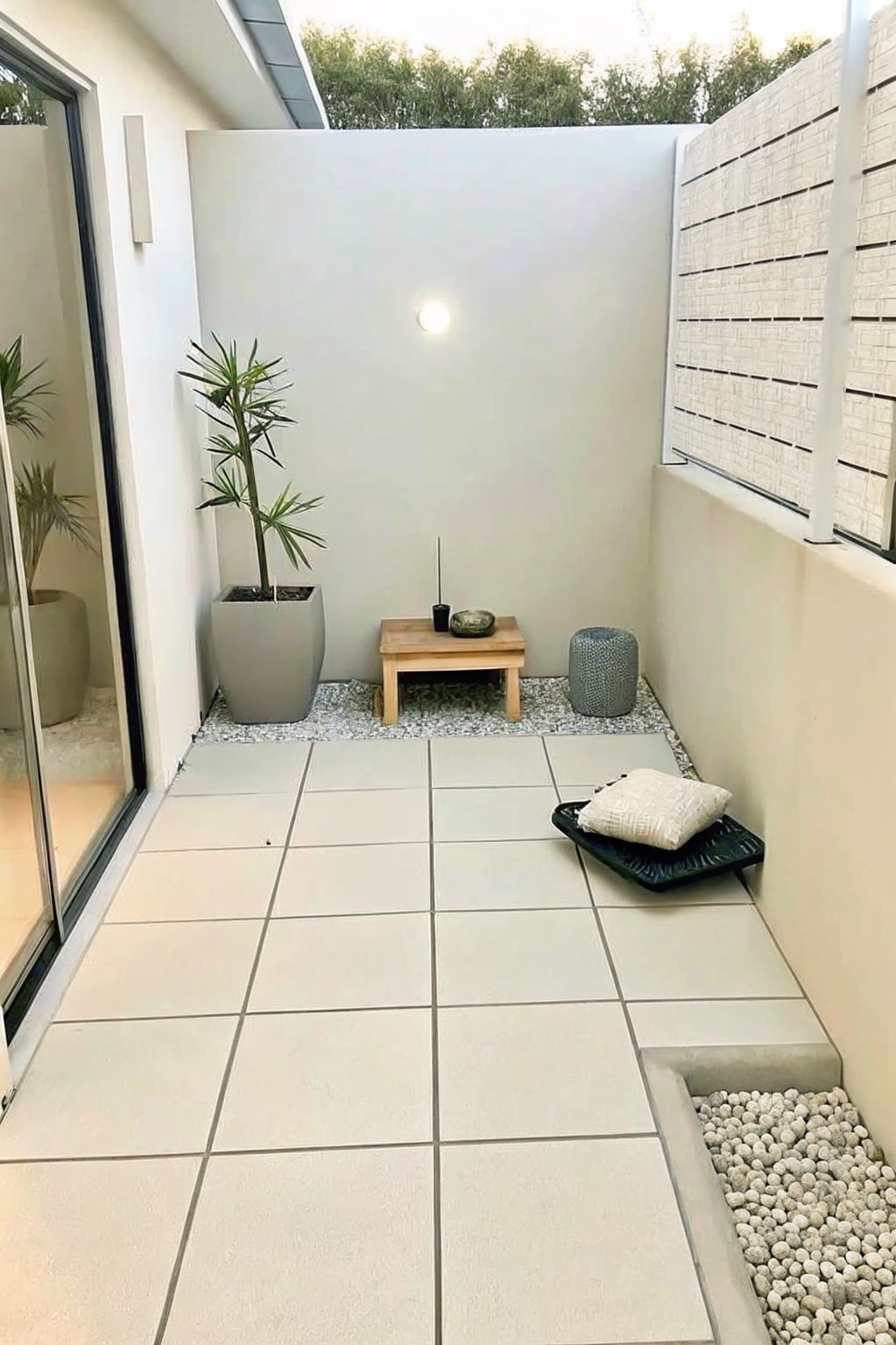 A small tiled patio with a low wooden table holding a bowl and candle, a large gray potted plant nearby, and a gravel border along the walls.