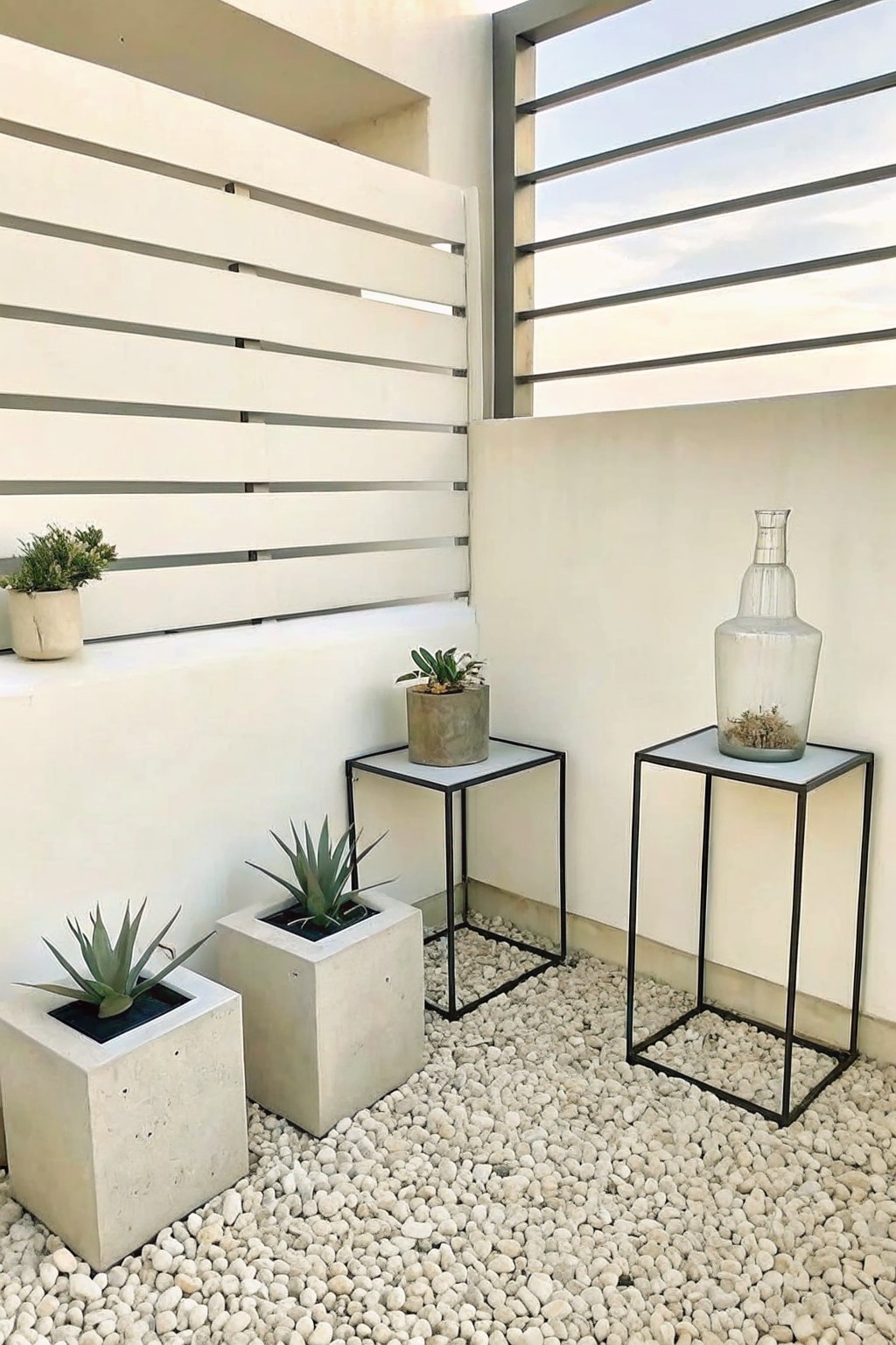 Outdoor patio with gravel floor, white walls, concrete block planters holding succulents, and small metal-framed tables displaying potted plants and a glass vase.