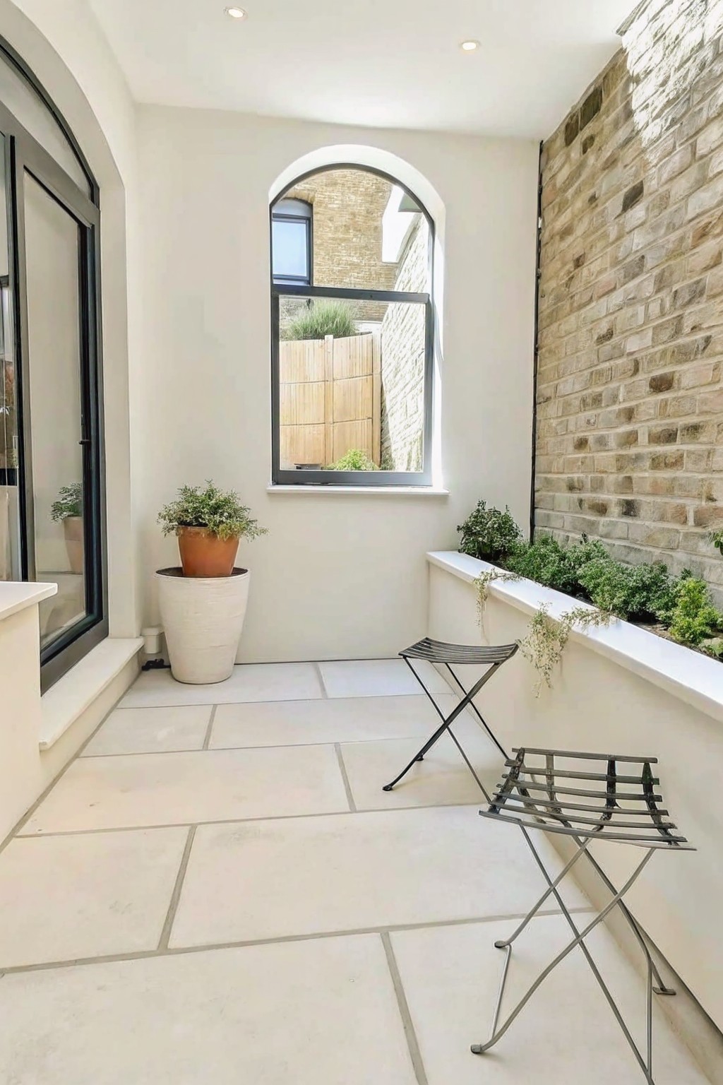 A compact outdoor patio with large light stone tiles, a tall brick wall on the right with a long built-in white stone planter ledge holding green plants, a large terracotta pot on the left, an arched window, and two metal folding stools.