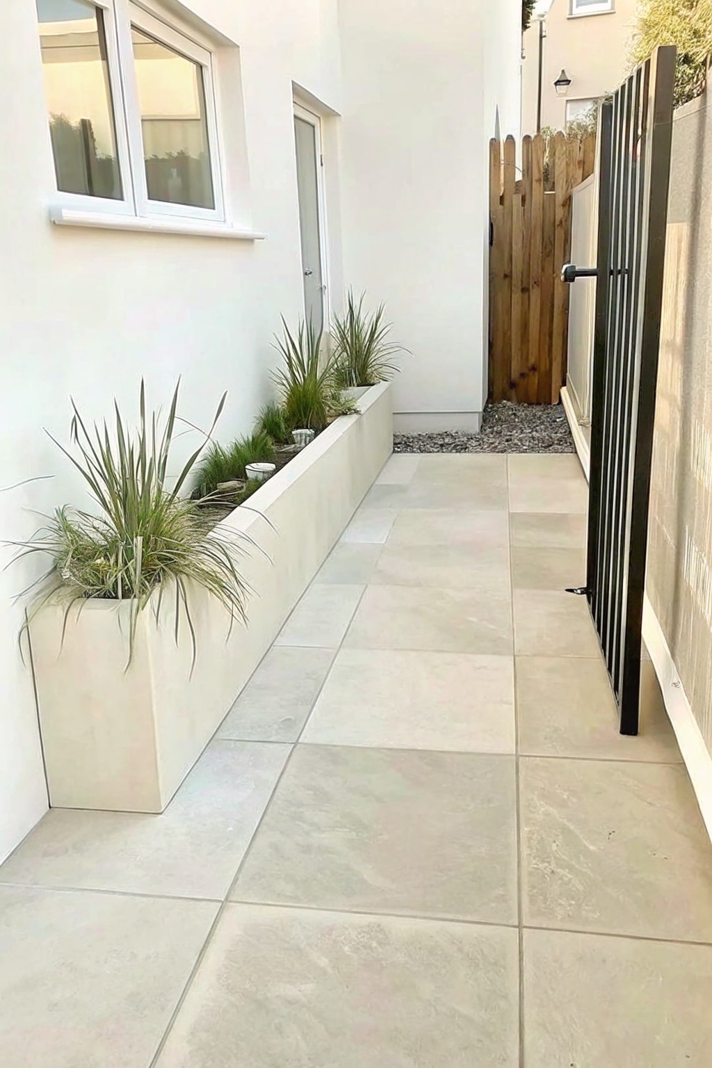 A narrow outdoor walkway with large light stone tiles, bordered by a long rectangular off-white planter filled with tall grasses along a white wall.