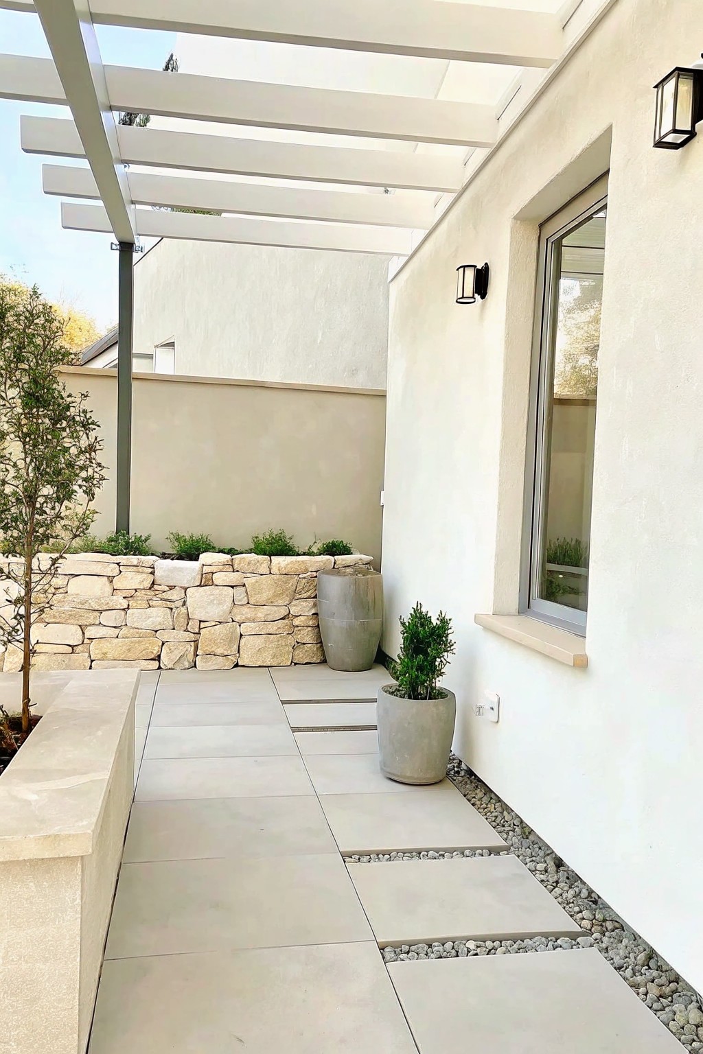 A paved outdoor patio with large cylindrical gray concrete planters, a low stone retaining wall, and light gray tiles under a white pergola.