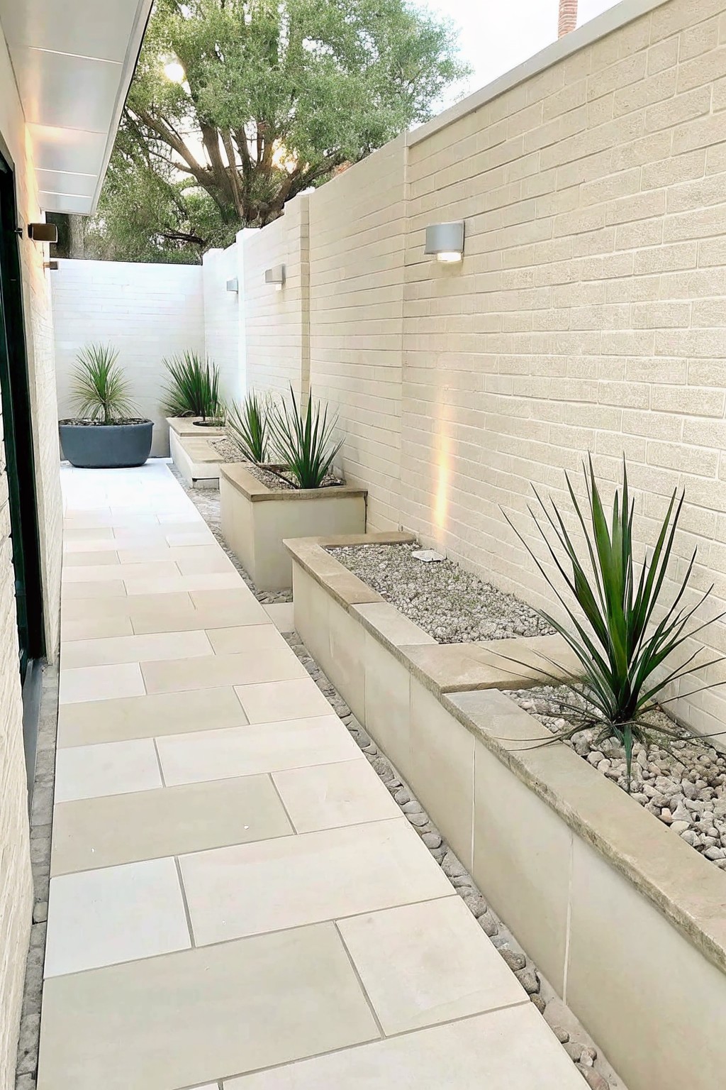 A narrow paved stone walkway runs beside a light brick wall lined with long rectangular concrete planters filled with gravel and spiky green plants.