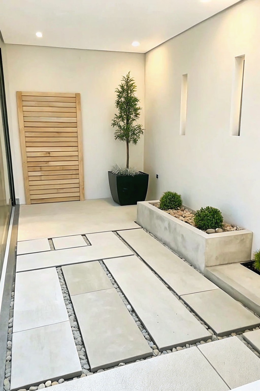 A modern outdoor entryway with large light gray stone pavers separated by white pebbles, a long raised concrete planter filled with shrubs and gravel, and a tall tree in a black pot beside a wooden slatted door.