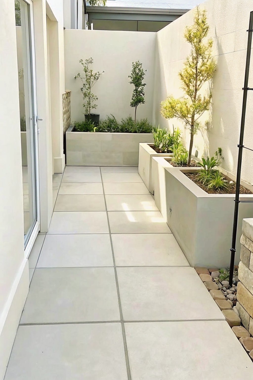 A narrow paved walkway lined on one side by rectangular light stone planters holding small trees and shrubs.