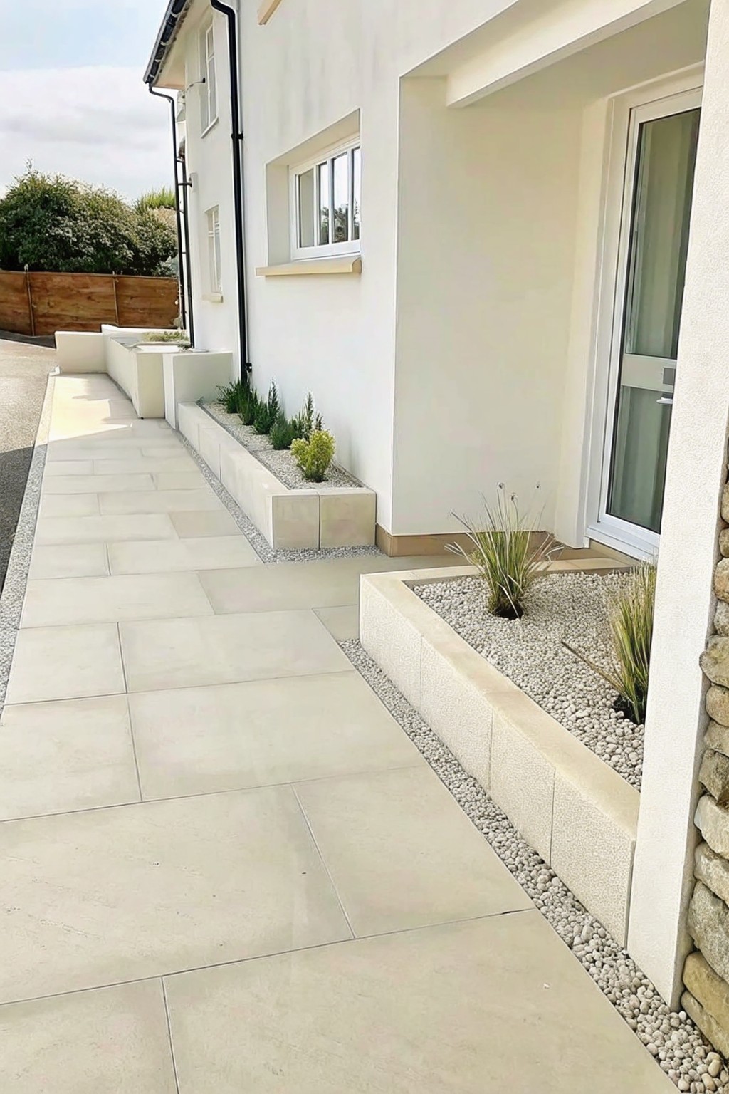A modern white house exterior with a light stone paved walkway bordered by raised rectangular concrete planters filled with gravel and small ornamental grasses.