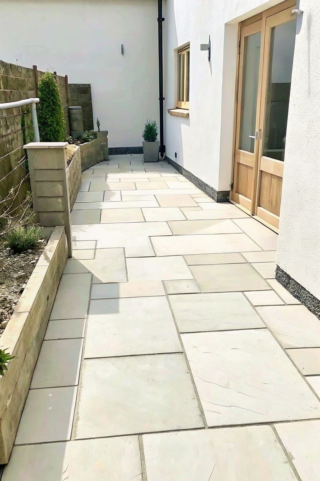 A narrow garden walkway made of large light stone pavers runs between a low stone retaining wall on the left and a white house wall with wooden doors on the right.