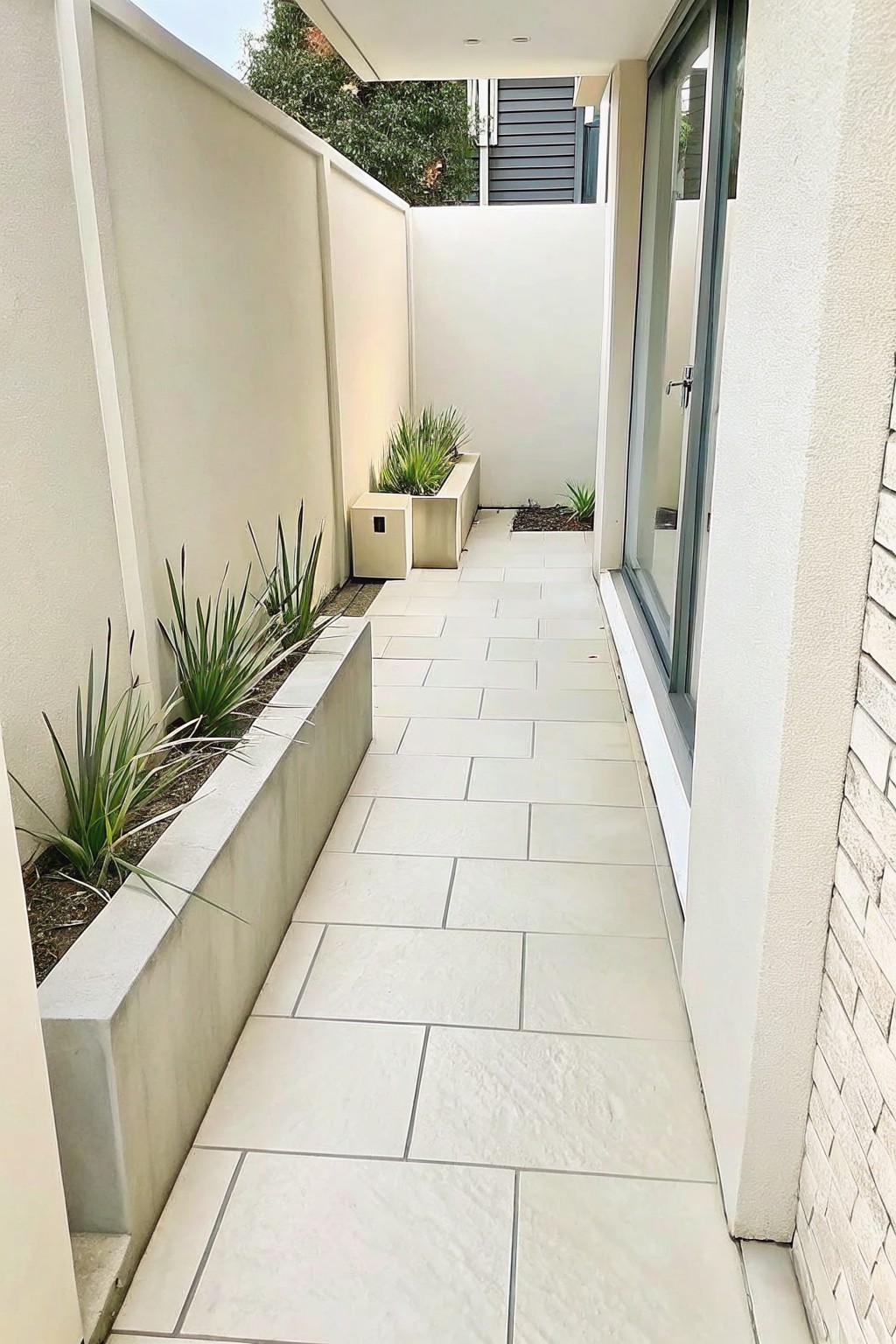 Narrow modern walkway with white tiles, beige walls, concrete planters of green plants, glass door.