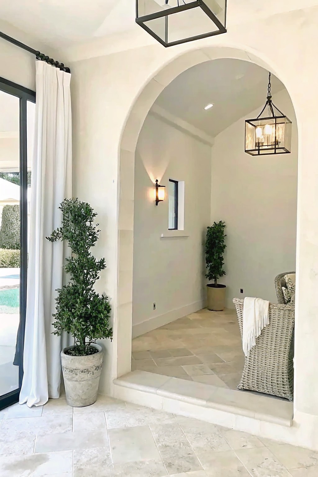 An interior view through a white arched opening shows a large stone planter with a tall shrub beside a glass door with a white curtain, and a matching planter visible outside on stone tile flooring.