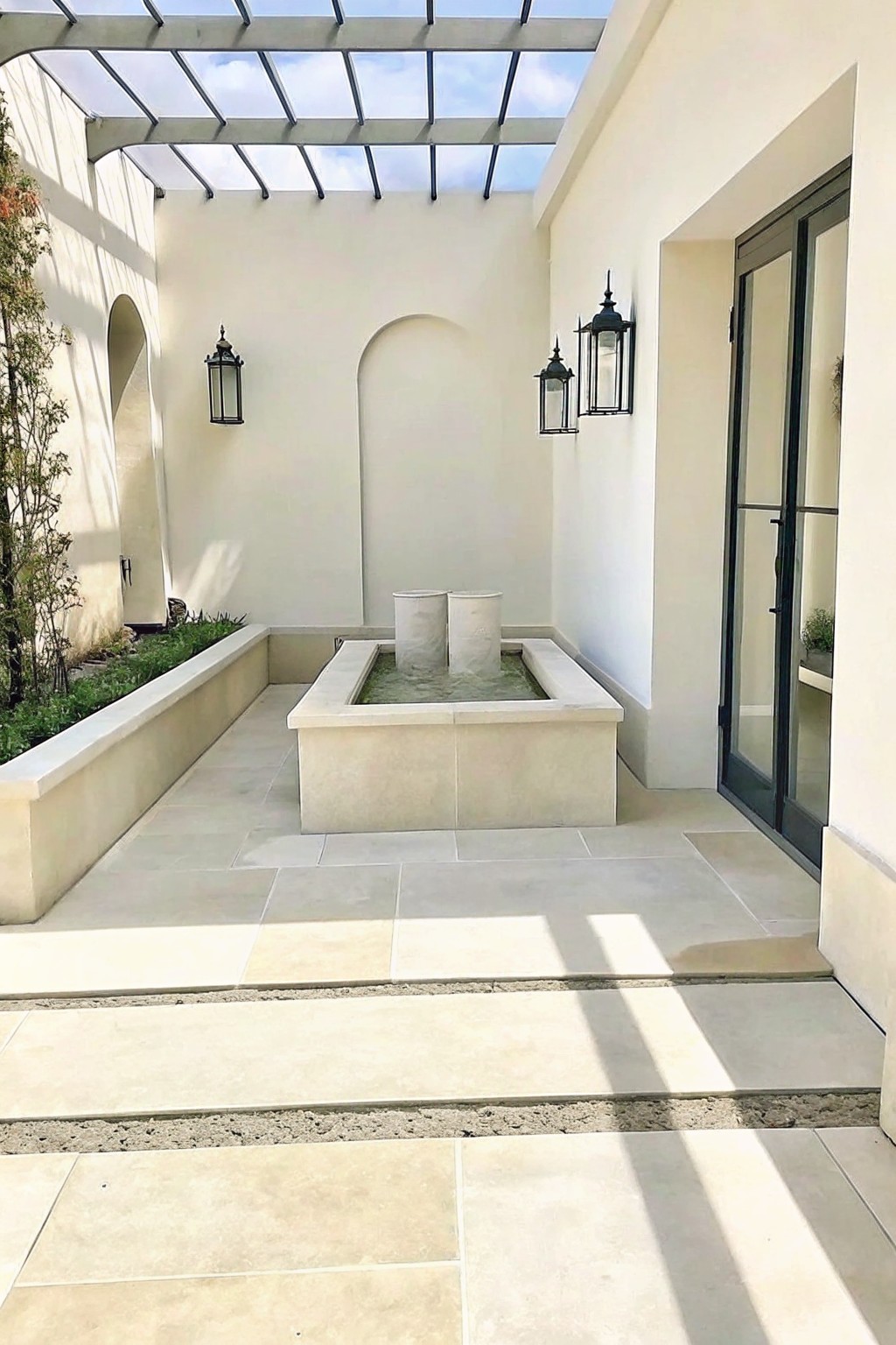 A square stone fountain basin in a light-tiled courtyard holds two short cylindrical stone planters, with white walls, black lanterns, and a glass pergola overhead.