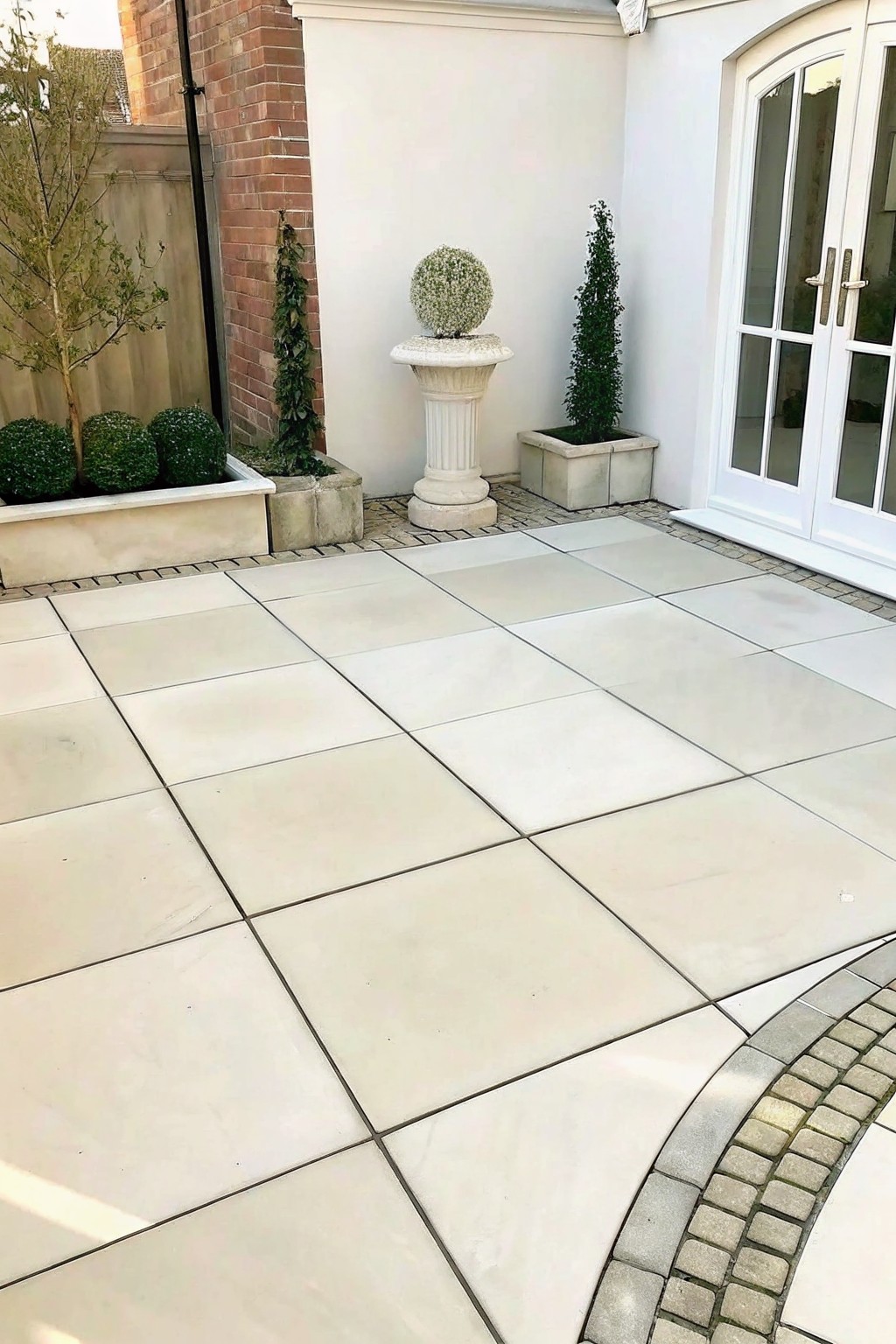 A paved courtyard with large light stone tiles, several rectangular stone planters holding trimmed shrubs, and a round stone pedestal holding a spherical plant.