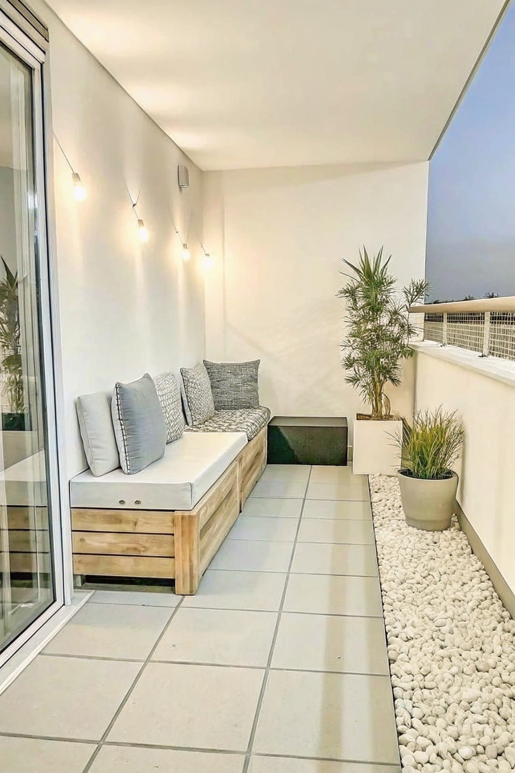 A narrow outdoor balcony with a built-in wooden bench along the left wall, white floor tiles, and a strip of white pebbles holding two potted plants on the right side.