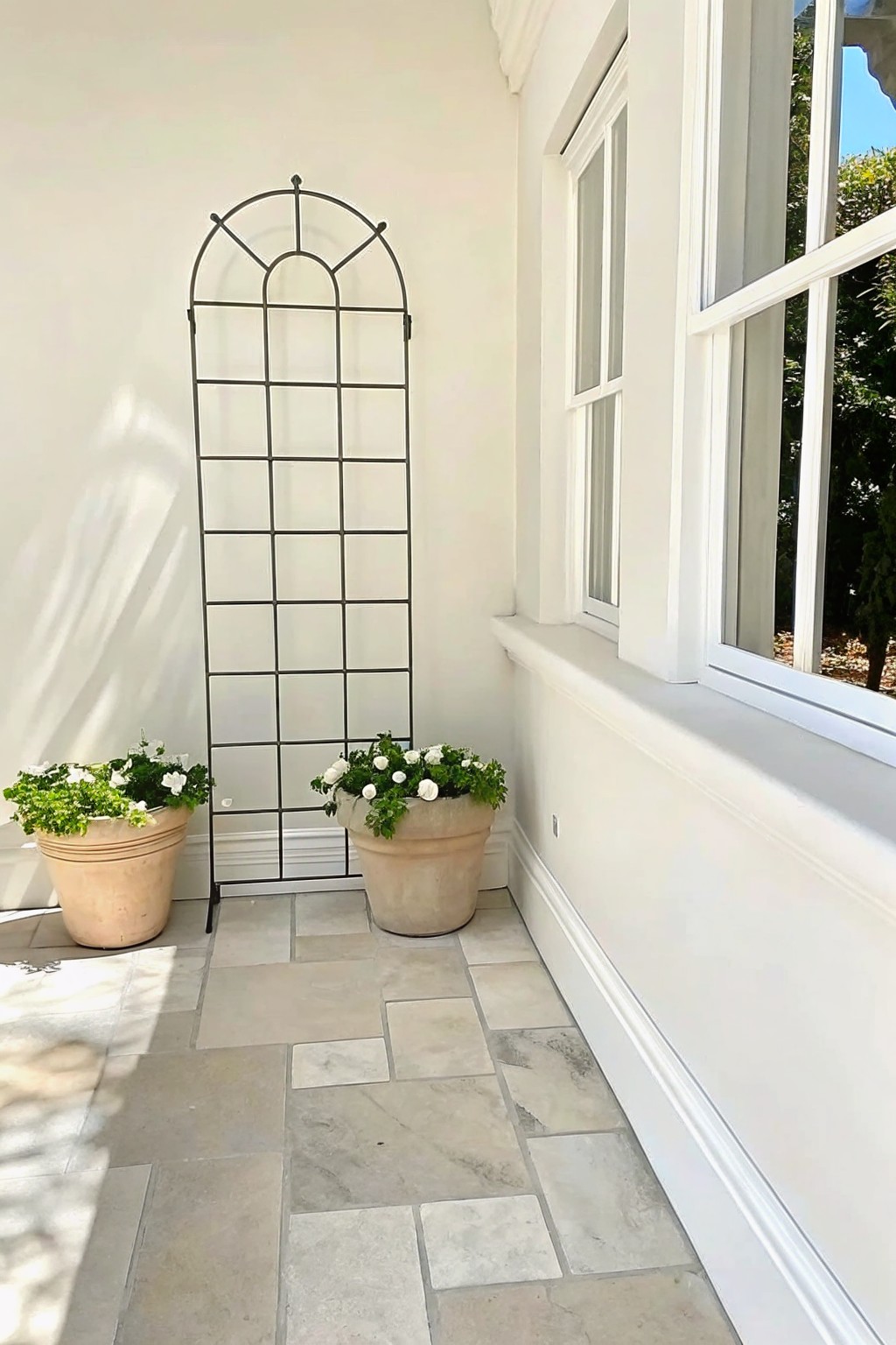 A covered porch with light stone tile flooring shows two beige stone planters holding white flowers placed on either side of a tall black metal arched trellis against a white wall next to a large window.